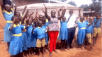 Group of children in blue uniforms with a teacher in a colorful top and red pants, standing on a dirt ground.