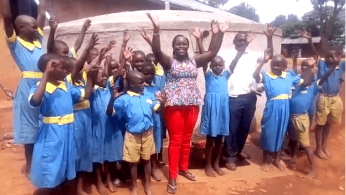 Group of children in blue uniforms with a teacher in a colorful top and red pants, standing on a dirt ground.